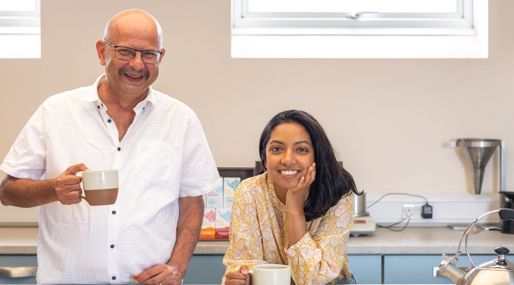Jimmy our tea blender who is an older south asian man holding a glass mug of Chai with Tina who is a young early 30 year old female of south asian heritage leaning on herleft hand with a big smile. 