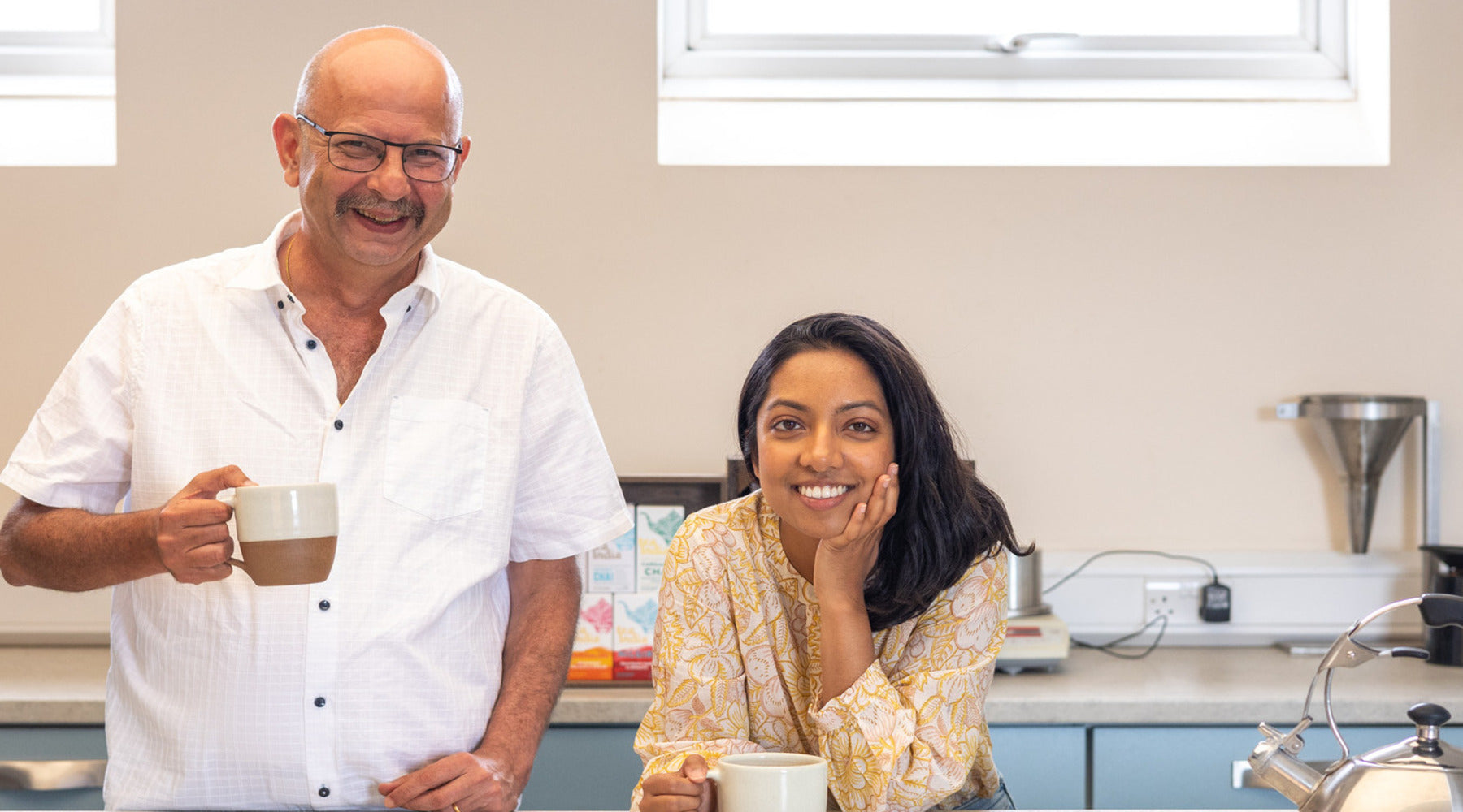 Jimmy our tea blender who is an older south asian man holding a glass mug of Chai with Tina who is a young early 30 year old female of south asian heritage leaning on herleft hand with a big smile.
